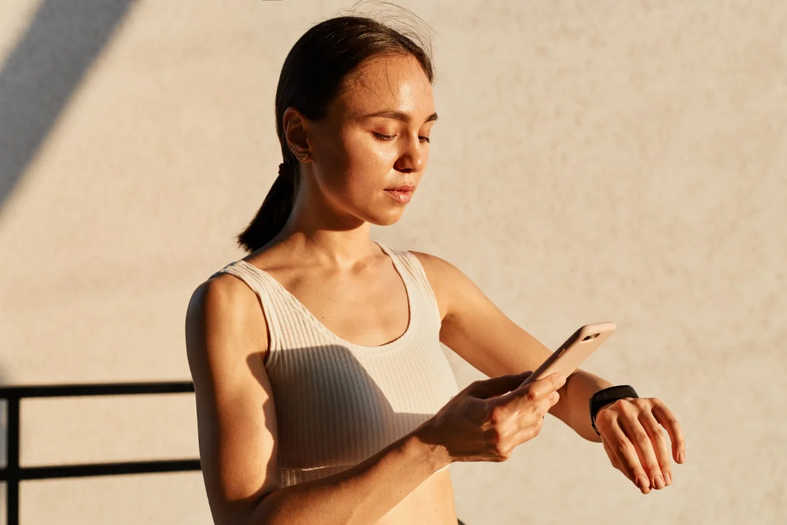 A woman in low sunlight checking her phone alongside a wrist tracker
